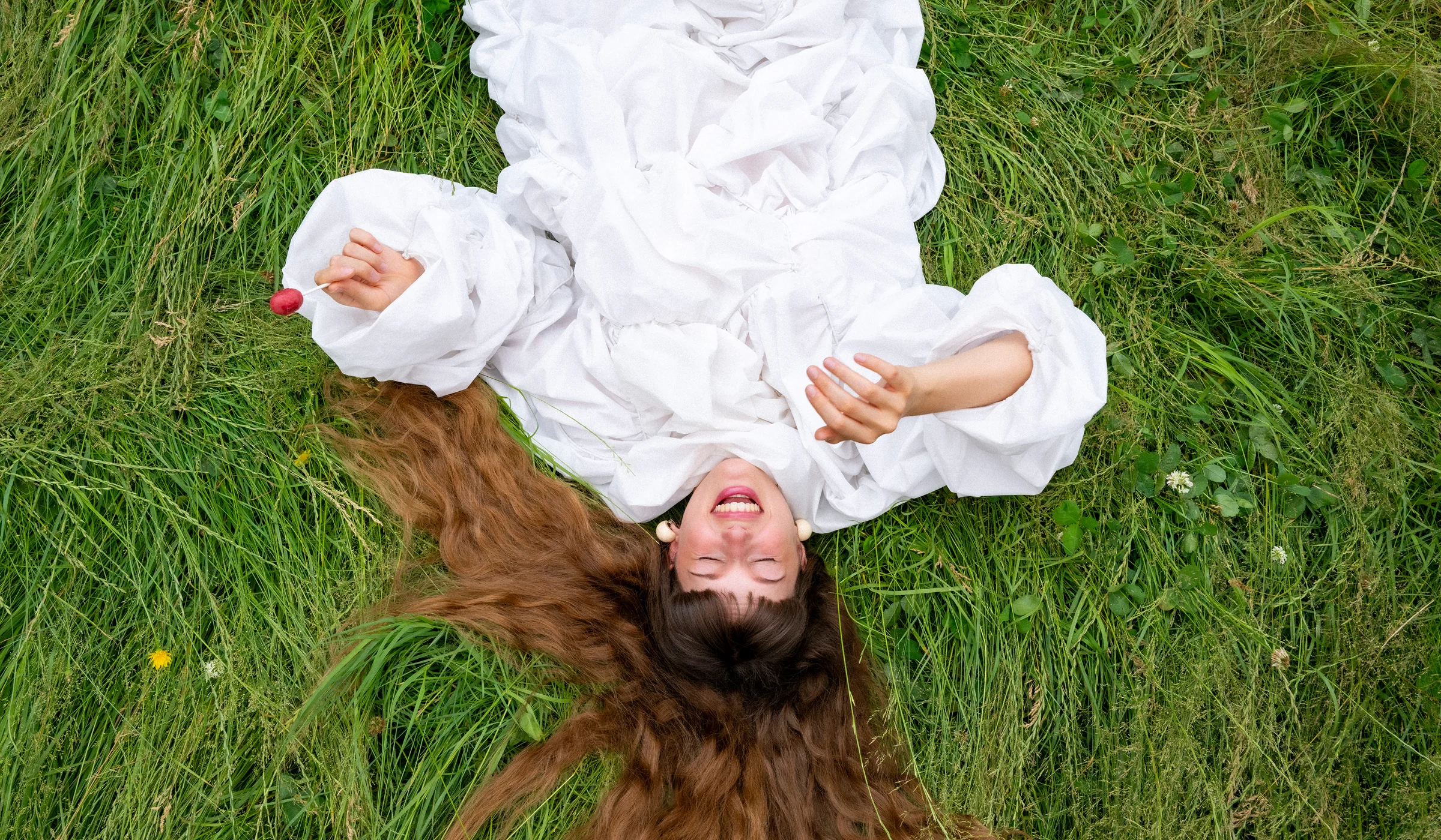 Top-view photograph of a smiling woman with long brown hair, dressed in white and holding a red lollipop while lying in green grass.